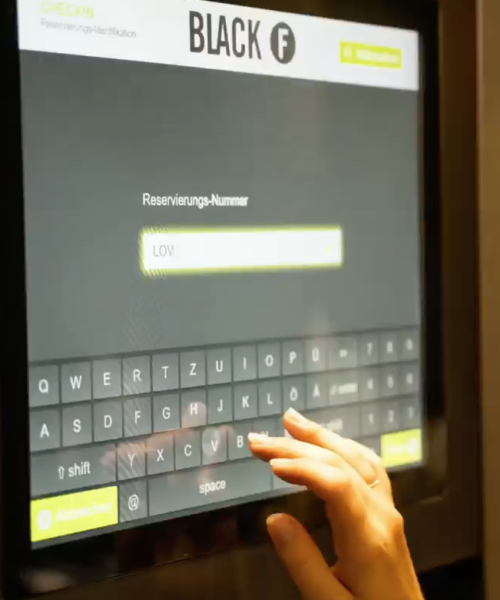 Checkin Bildschirm BlackF Die Hand einer Frau bedient den Touchscreen des digitalen Self-Check-in-Terminals von BLACKF und gibt die Reservierungsnummer ein.