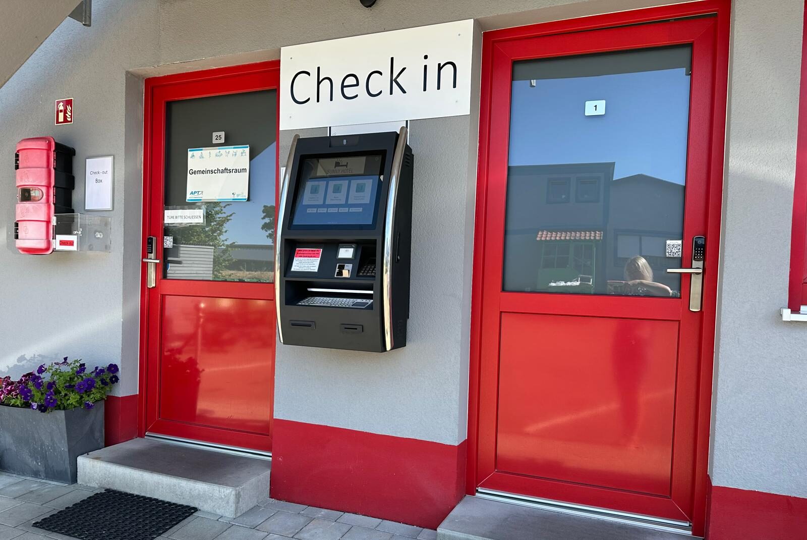 Self-Check-in-Terminal zwischen zwei Zimmertüren im Sunny Hotel Regenstauf mit rotem Eingangsbereich